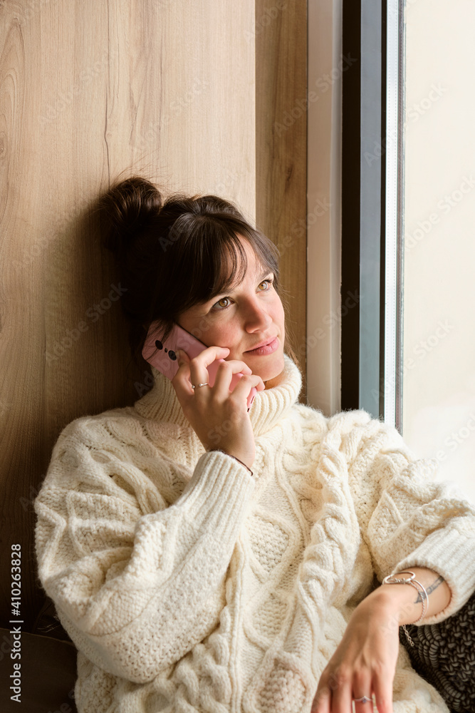 Beautiful woman in sweater looking up while talking on mobile phone by window at home Beautiful woman in sweater looking up while talking on mobile phone by window at home