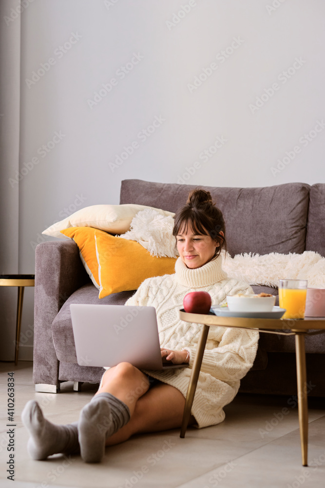 Woman in sweater using laptop while sitting on floor in living room at home Woman in sweater using laptop while sitting on floor in living room at home