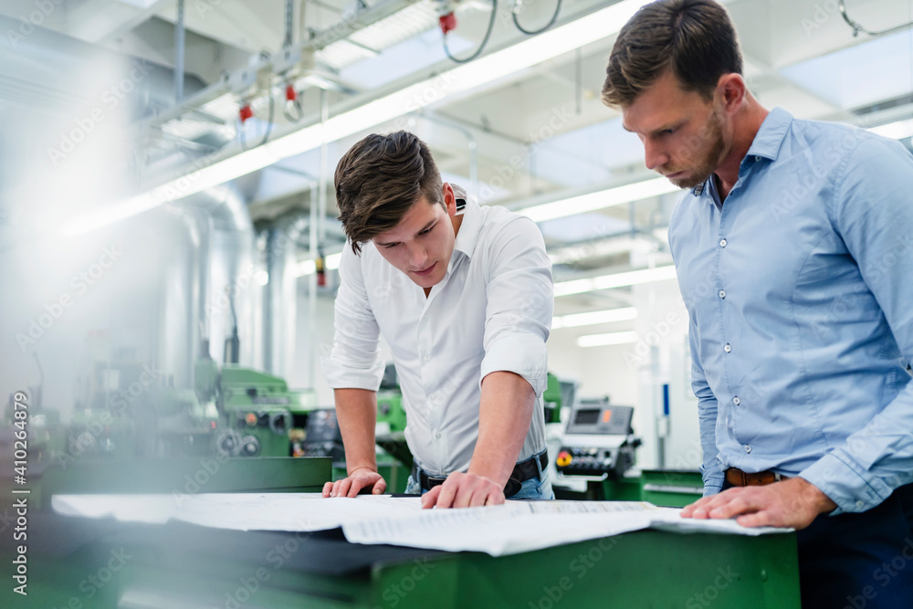 Male entrepreneur with colleague looking at blueprint on desk in factory Male entrepreneur with colleague looking at blueprint on desk in factory