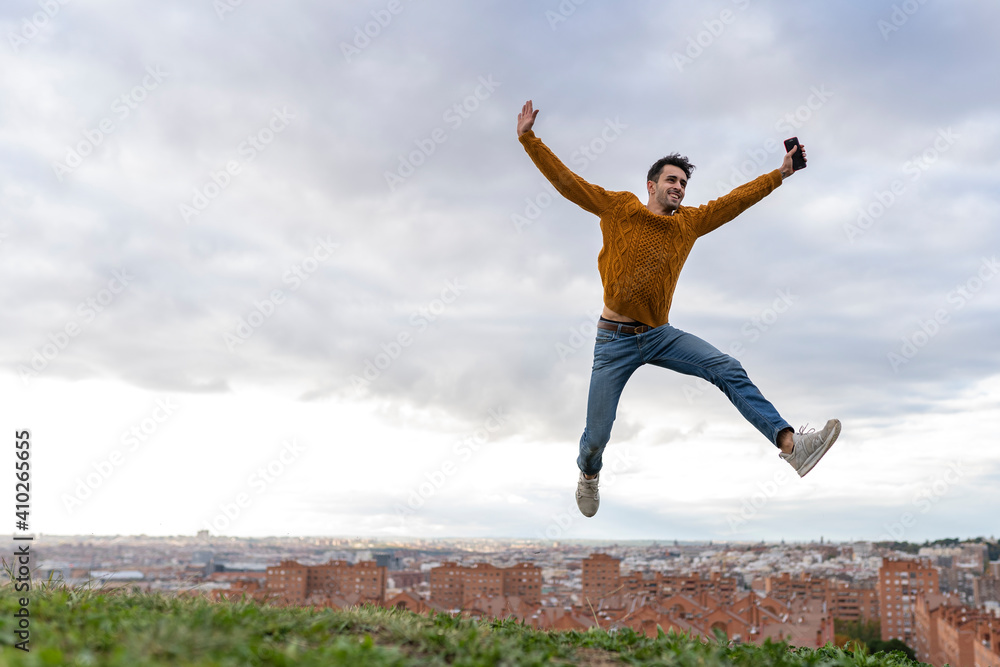 Excited young man holding mobile phone while jumping on hill against cityscape Excited young man holding mobile phone while jumping on hill against cityscape