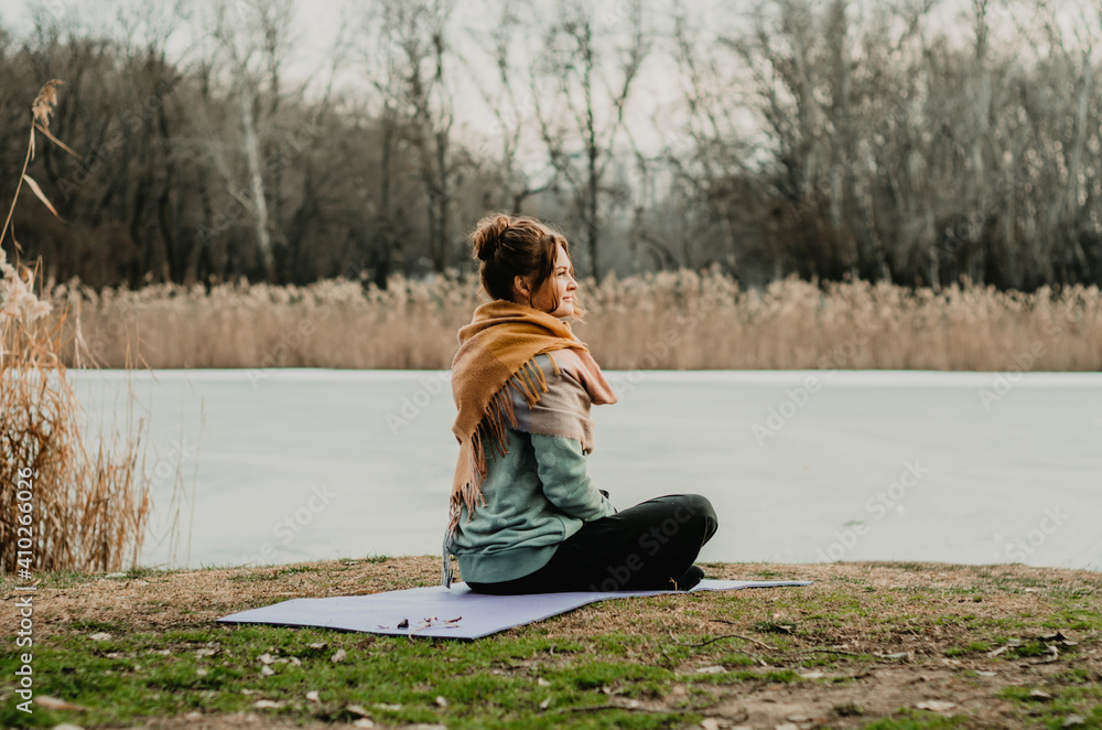 Fototapeta premium Back view of young woman in comfy clothes seated in meditation on the ground in a park, with a lake background. Concept: self care practices, movement, introspection