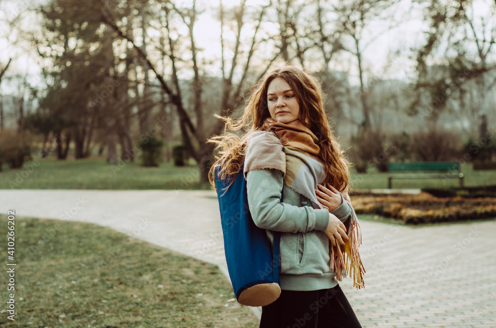 Portrait of a young woman with long ginger hair in motion, walking in the park with a yoga mat on the shoulder Portrait of a young woman with long ginger hair in motion, walking in the park with a yoga mat on the shoulder