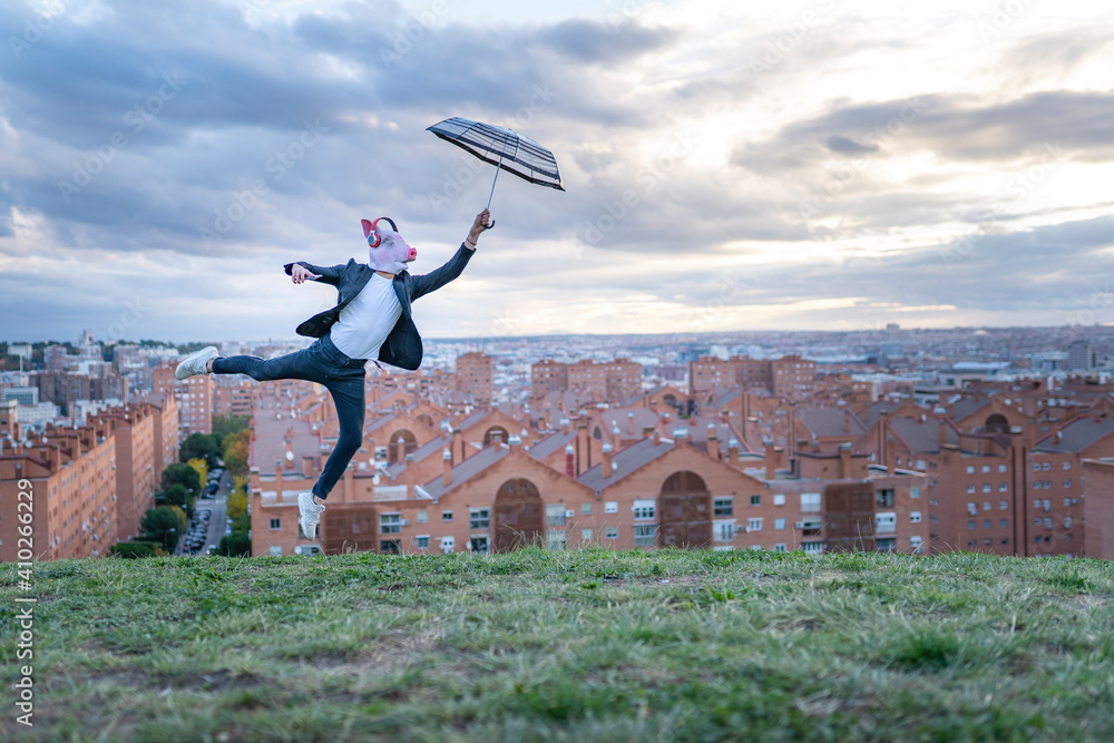Male professional wearing pig mask holding umbrella while jumping on hill at sunset Male professional wearing pig mask holding umbrella while jumping on hill at sunset