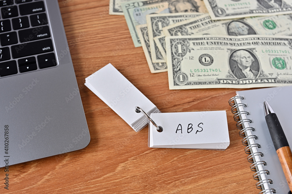 On the desk there are bills, a laptop, and a word book with the word ABS written on it. It was an abbreviation for the financial term Asset Backed Security. On the desk there are bills, a laptop, and a word book with the word ABS written on it. It was an abbreviation for the financial term Asset Backed Security.