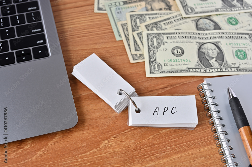 On the desk there are bills, a laptop, and a word book with the word APC written on it. It was an abbreviation for the financial term average propensity to consume. On the desk there are bills, a laptop, and a word book with the word APC written on it. It was an abbreviation for the financial term average propensity to consume.
