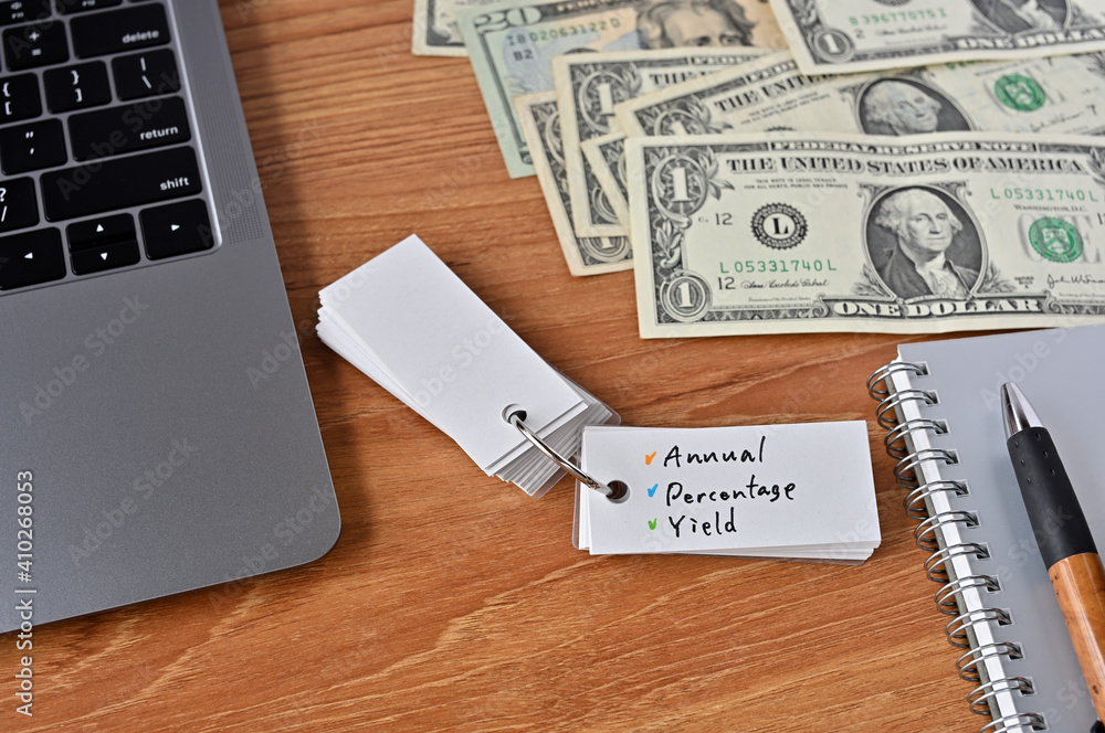 On the desk there were bills, a laptop, and a word book with the word annual percentage yield written on it. On the desk there were bills, a laptop, and a word book with the word annual percentage yield written on it.