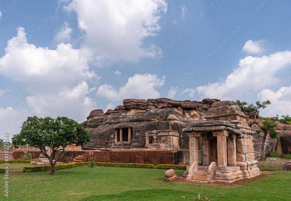 Aihole, Karnataka, India - November 7, 2013: Ravanaphadi Cave Shiva Temple. Green landscape with brown stone temple buiidings and black rock cave under blue cloudscape.