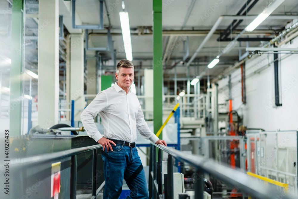Smiling mature businessman with hand on hip standing by railing in factory Smiling mature businessman with hand on hip standing by railing in factory