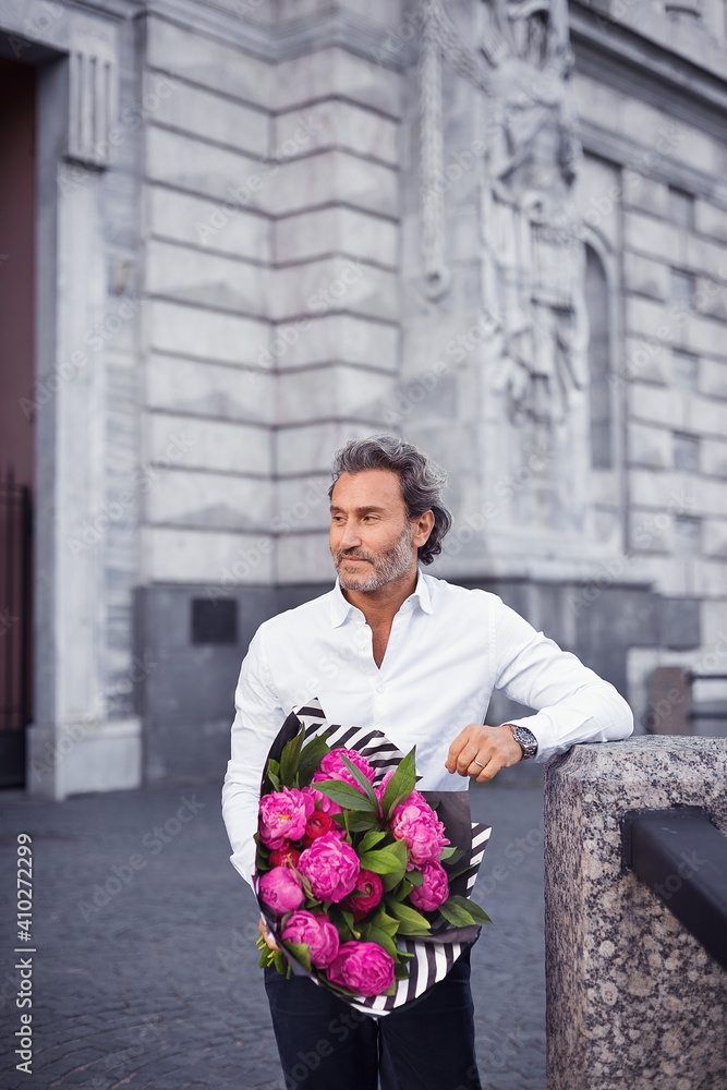Fototapeta premium A handsome stylish man in a white shirt and black trousers and a bouquet of pink peonies in his hands waiting for his beloved girl.
