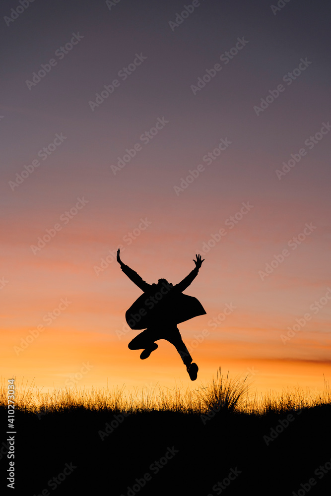 Carefree man wearing jacket jumping with arms outstretched against sky during sunset Carefree man wearing jacket jumping with arms outstretched against sky during sunset