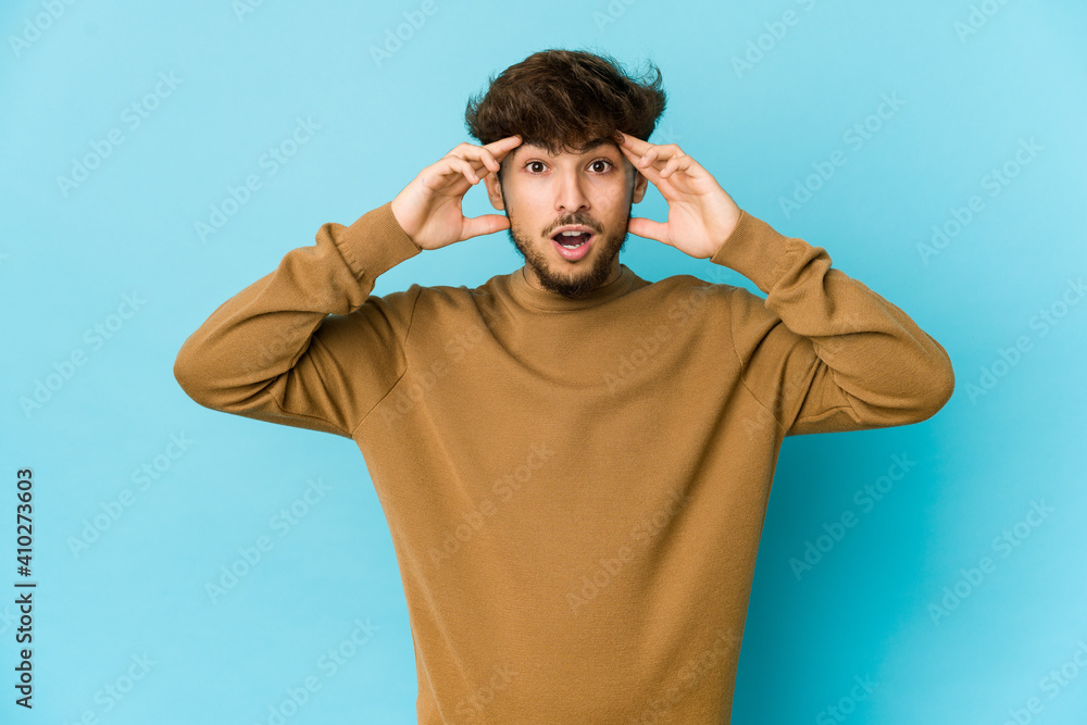 Naklejka premium Young arab man on blue background receiving a pleasant surprise, excited and raising hands.