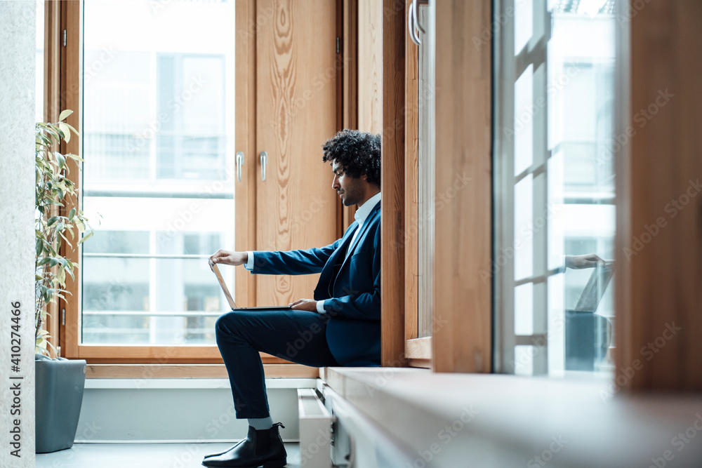 Young male entrepreneur using laptop while sitting against window at workplace Young male entrepreneur using laptop while sitting against window at workplace