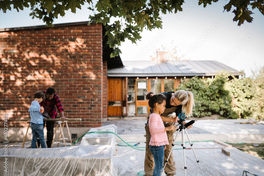 Mother talking to daughter by surveyor while son learning carpentry from father outside house Mother talking to daughter by surveyor while son learning carpentry from father outside house