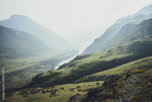 Misty mountain landscape with hills and rocks on background of wide mountain river in mist. Atmospheric scenery with mountain relief and big river in dark green valley in rainy weather. Gloomy weather