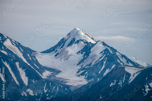 Awesome mountains landscape with big snowy mountain pinnacle in blue white colors under cloudy sky. Atmospheric highland scenery with high mountain wall with pointed top with snow under white clouds. © Daniil