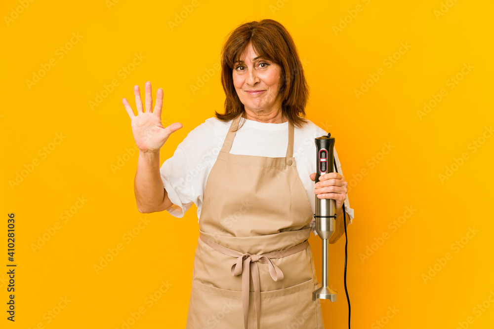 Middle age caucasian cook woman holding a mixer isolated smiling cheerful showing number five with fingers.