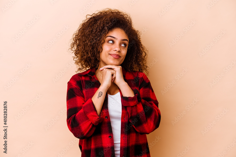 Young african american woman isolated on beige background keeps hands under chin, is looking happily aside. Young african american woman isolated on beige background keeps hands under chin, is looking happily aside.