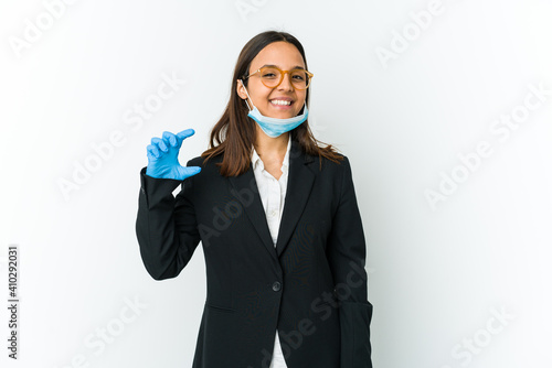Young business latin woman wearing a mask to protect from covid isolated on white background holding something little with forefingers, smiling and confident.