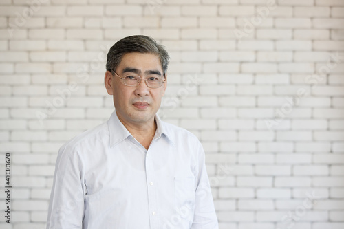 Portrait of a happy Asian senior businessman wearing glasses on white brick wall background and looking at camera with kindly smile. Close up face of happy successful business man