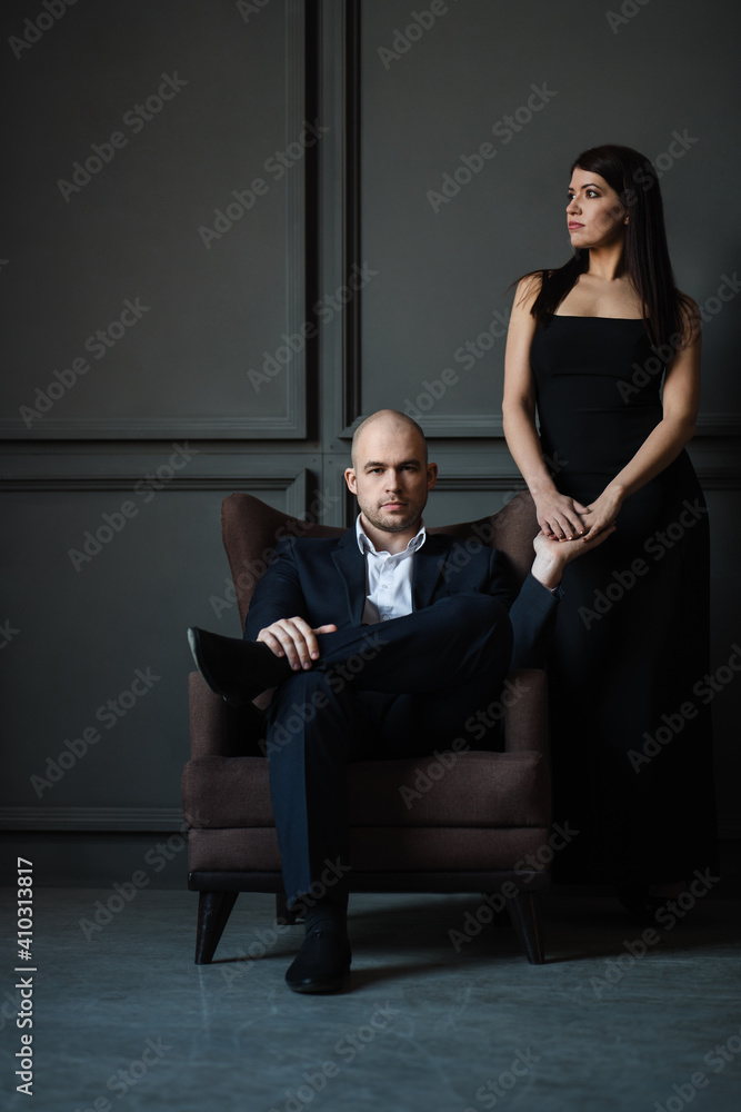 Elegant couple in love. A man and a woman in a dark room. The girl in the black dress is standing and the bald guy in a suit sitting in a chair, loft. Vertical photo. Valentine's Day Elegant couple in love. A man and a woman in a dark room. The girl in the black dress is standing and the bald guy in a suit sitting in a chair, loft. Vertical photo. Valentine's Day