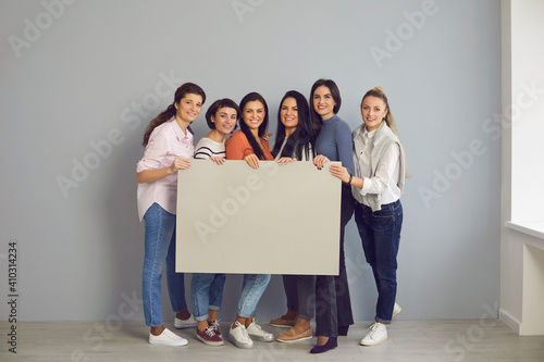 Group of happy young women in their 20s and 30s standing together in office or studio, smiling at camera and holding empty gray cardboard sign, mock-up banner, grey blank poster with space for text photo