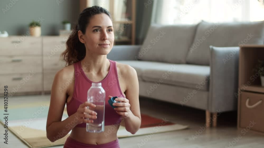 Waist-up shot of brown-eyed sporty Caucasian young woman with ponytail hairstyle wearing crop top, sitting in living room, drinking water from bottle and smiling Waist-up shot of brown-eyed sporty Caucasian young woman with ponytail hairstyle wearing crop top, sitting in living room, drinking water from bottle and smiling