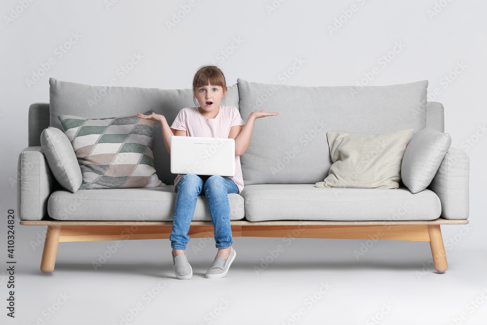 Surprised little girl with laptop on sofa against light background