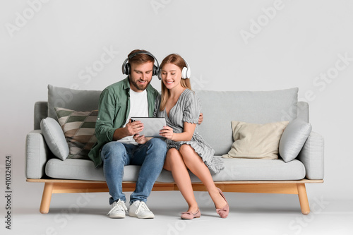 Young couple with tablet computer and headphones on sofa against light background