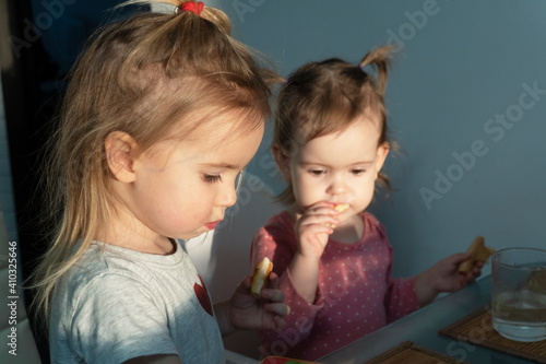 Two cute little caucasian girls sitting at the table  eating  looking. Sun shines at the girls  happy childhood and family concept.