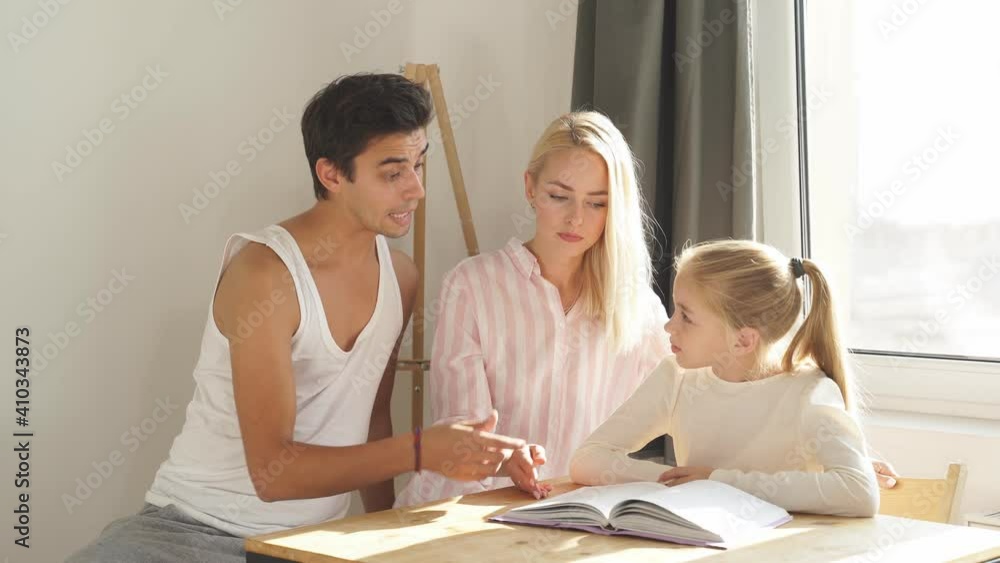 Young white family busy together in their kitchen. Young parents helping cute school child girl doing homework sitting at kitchen table. Young white family busy together in their kitchen. Young parents helping cute school child girl doing homework sitting at kitchen table.