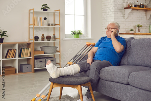 Injured older adult spends recovery period at home: Senior man with broken leg in plaster cast sitting on sofa with mobile phone, sharing news with family or talking to doctor about his bone fracture photo