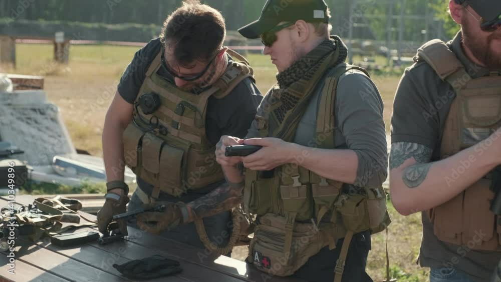 Medium handheld shot of team of three tattooed caucasian men in military uniform assembling guns outdoors on sunny day Medium handheld shot of team of three tattooed caucasian men in military uniform assembling guns outdoors on sunny day