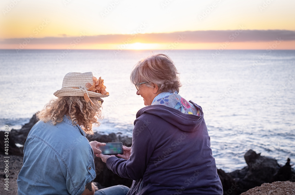 Fototapeta premium Two women sitting admiring the sunset over the sea while using their mobile phone. An elderly person and a curly middle-aged people enjoy freedom and holiday