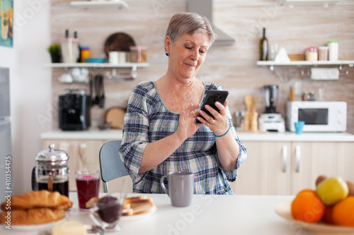 Relaxed elderly woman browsing on phone in kitchen during breakfast. Grandma using modern smartphone internet technology, online communication connected to the world, senior leisure time with gadget