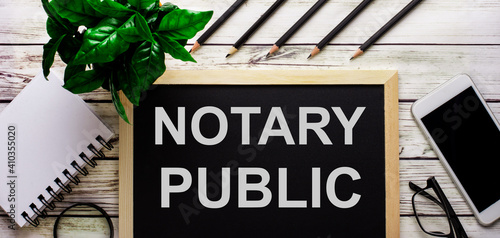 NOTARY PUBLIC is written in white on a black board next to a phone, notepad, glasses, pencils and a green plant. photo