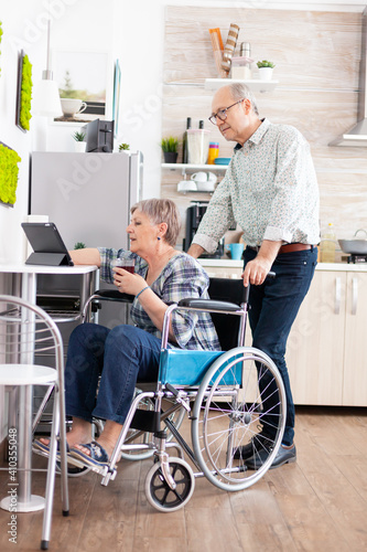 Paralysied senior wife in wheelchair working at tablet computer, showing to husband her project sitting in kitchen. Disabled handicapped old elderly person using modern communication online internet