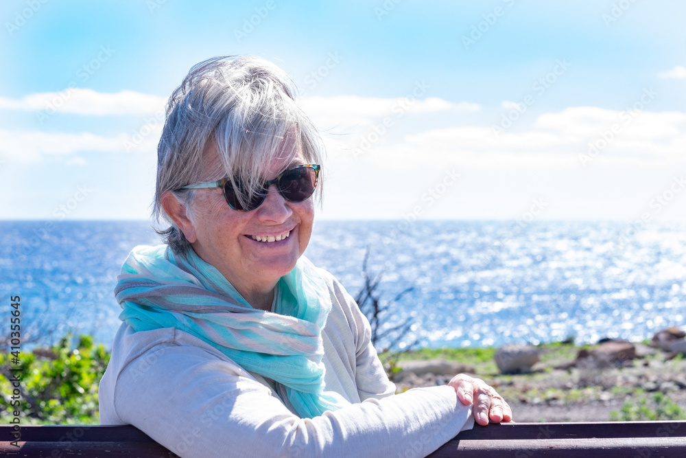 Portrait of a senior smiling woman relaxing in front to the sea, sitting on a bench looking at the camera. Senior woman enjoying retirement and outdoors