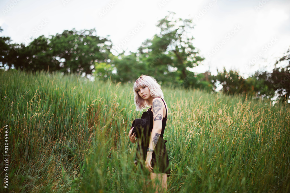 Young stylish girl with blonde hair standing in the green field in black dress, holding black hat. Portrait shot Young stylish girl with blonde hair standing in the green field in black dress, holding black hat. Portrait shot