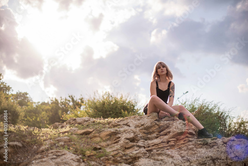Young stylish blonde woman with tattoos in black dress sitting on a big rock outdoors on sunny day