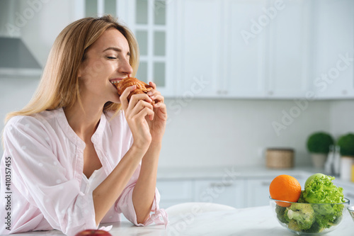 Concept of choice between healthy and junk food. Woman eating croissant at white table in kitchen