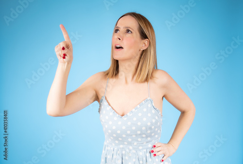 Portrait of a happy young woman wearing casual dress pointing finger up at copy space isolated over blue background