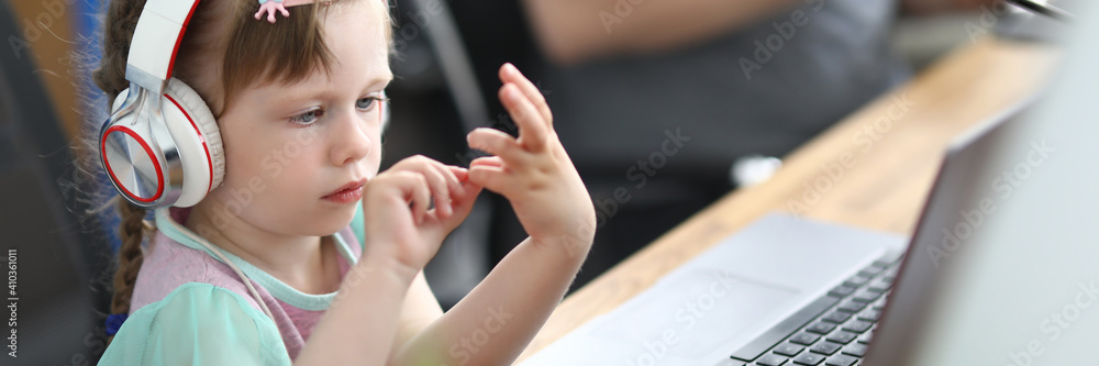 Portrait of cute little child sitting in parents office. Serious kid wearing white headset and listening music. Modern laptop on wooden desk. Business and technology concept Portrait of cute little child sitting in parents office. Serious kid wearing white headset and listening music. Modern laptop on wooden desk. Business and technology concept