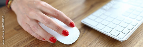 Close-up of person working on computer. Woman with red nails holding mouse for work. White keyboard on wooden desktop. Mobile phone on table. Business and career concept