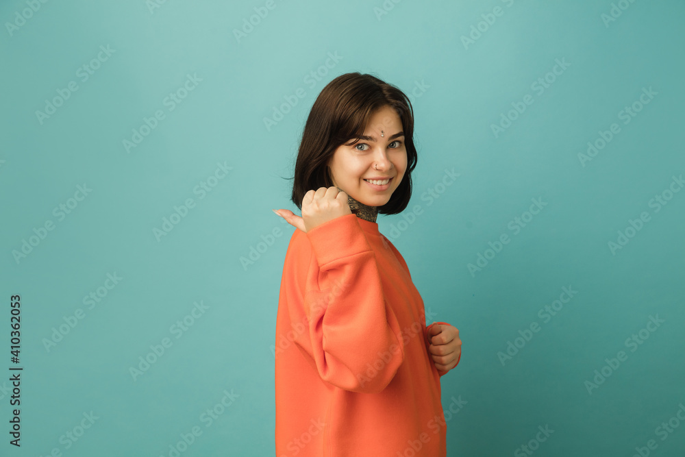 Fototapeta premium Pointing at side. Caucasian woman's portrait isolated on blue background with copyspace. Beautiful female model in orange hoodie. Concept of human emotions, facial expression, sales, ad, fashion.