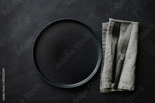 Empty black plate with black cutlery and gray linen napkin on black stone table. Top view.