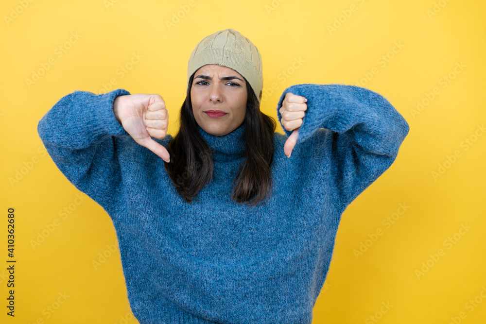 Young beautiful woman wearing blue casual sweater and wool hat with angry face, negative sign showing dislike with thumbs down Young beautiful woman wearing blue casual sweater and wool hat with angry face, negative sign showing dislike with thumbs down