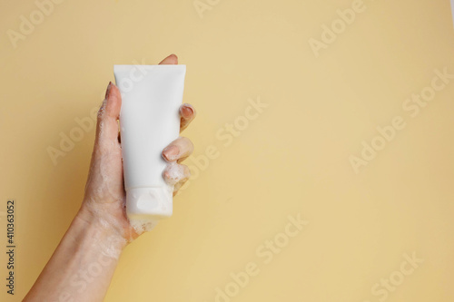 Fotografía female hand with soapy foam holds cosmetic tube on light yellow background