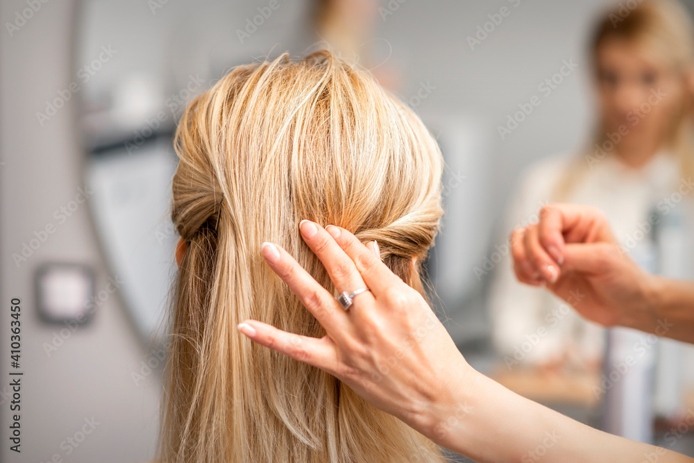 Fototapeta premium Back view of the female hand of hairdresser models a hairstyle of a young blonde woman in a hair salon