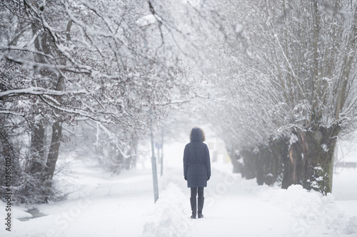 Young woman slowly walking on snow covered sidewalk through alley of trees in white snowy winter day at park after blizzard. Foggy air. Spending time alone in nature. Peaceful atmosphere. Back view. © fotoduets