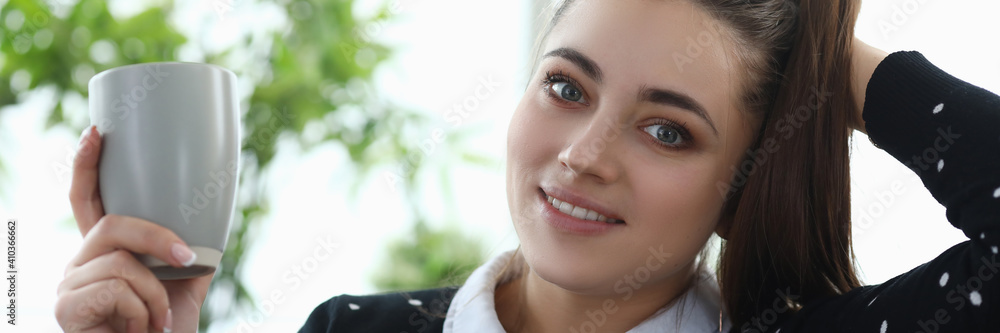 Fototapeta premium Portrait of lovely young woman drinking beverage from grey plastic cup. Brunette attractive female resting on cozy couch at home. Spare time and relaxation concept